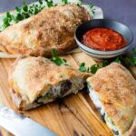 Two turkey, mushroom and broccoli calzones on a wooden cutting board with marinara dipping sauce and a knife.