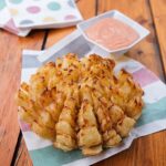 Blooming onion on a colored napkin on a wooden table with a dipping sauce near by.