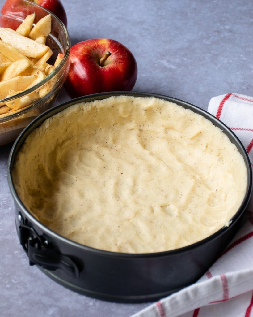 The crust for apple almond torte pressed into the bottom of a springform pan with a bowl of sliced apples in the background.