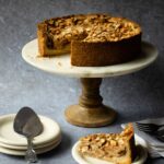Apple Almond Torte on a cake stand with a piece cut out on a white plate below.