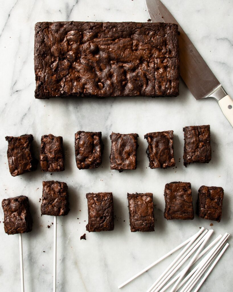 Brownies being cut into little rectangles on a marble surface with lollipop sticks and a knife.