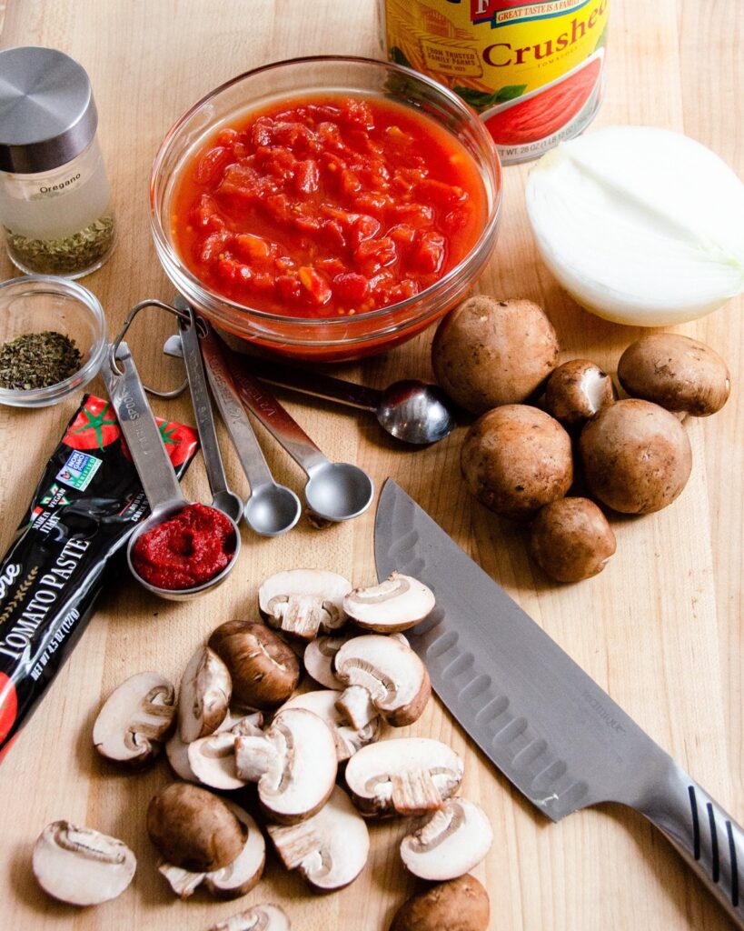 Ingredients on a wooden cutting board with  a  knife - tomatoes, mushrooms, tomato paste, herbs and half an onion.