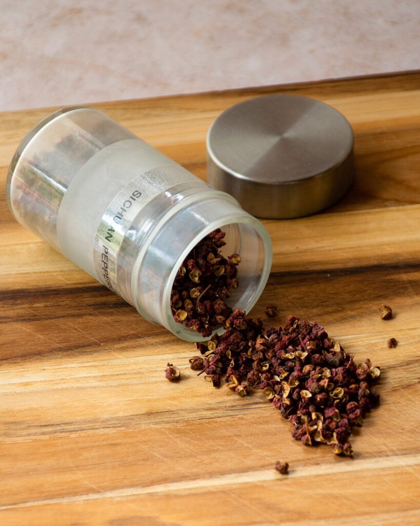 Sichuan Peppercorns tumbling out of a spice jar onto a cutting board.