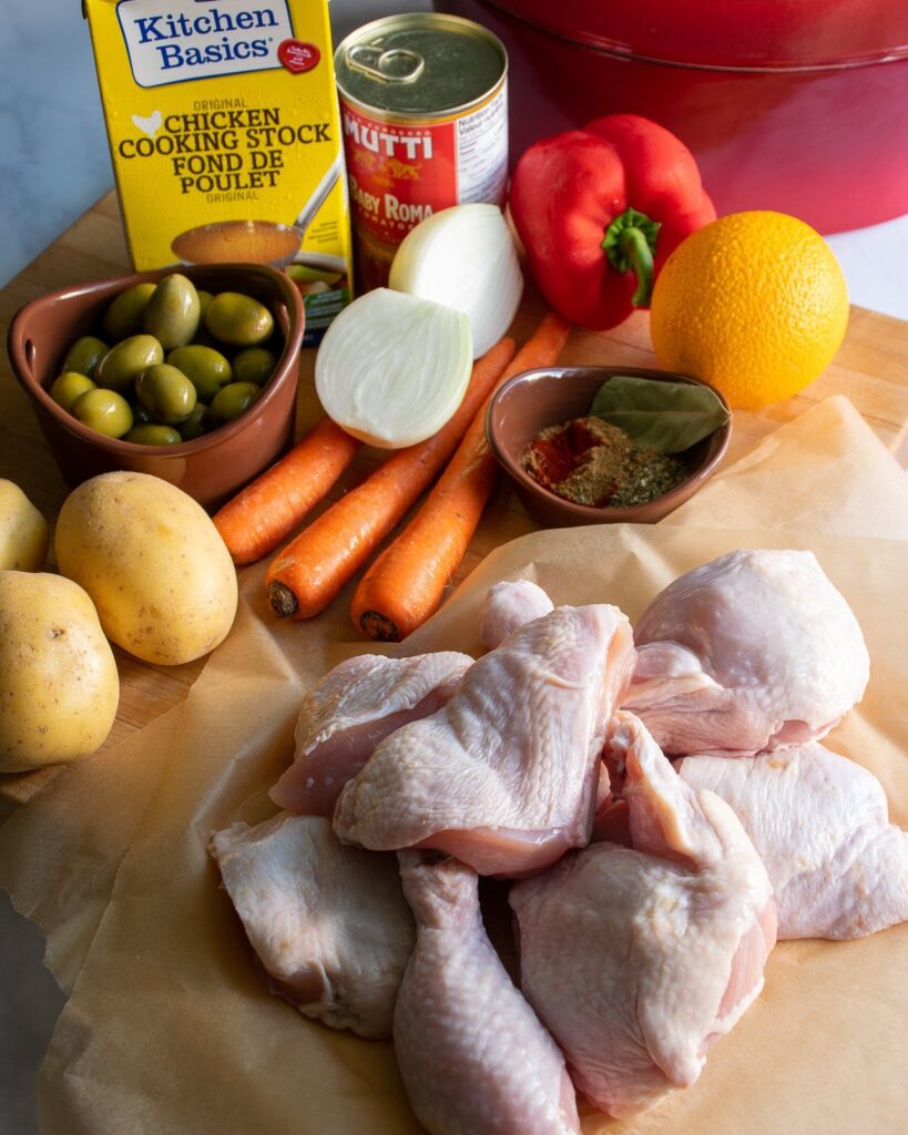 Raw ingredients on a cutting board - chicken, potatoes, carrots, olives, onion, red pepper, orange, spices, chicken stock and a can of tomatoes.