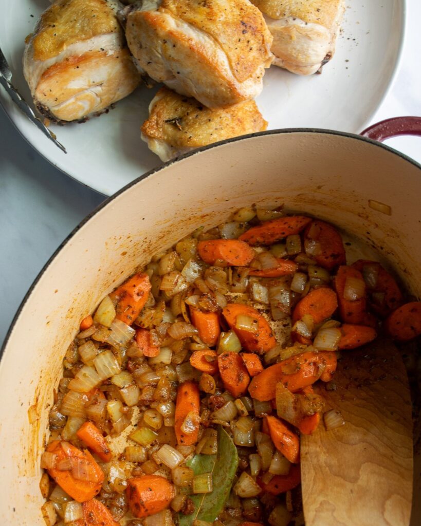 A Dutch oven with sautéed vegetables and spices with a plate of browned chicken beside it.
