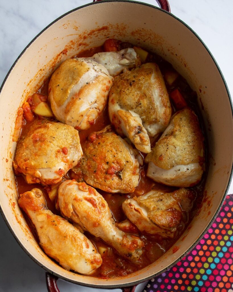 A Dutch oven with chicken pieces and a tomato based braising liquid.