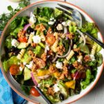 Tuna Greek Salad in a white bowl on a white wooden table with dressing and a blue napkin.