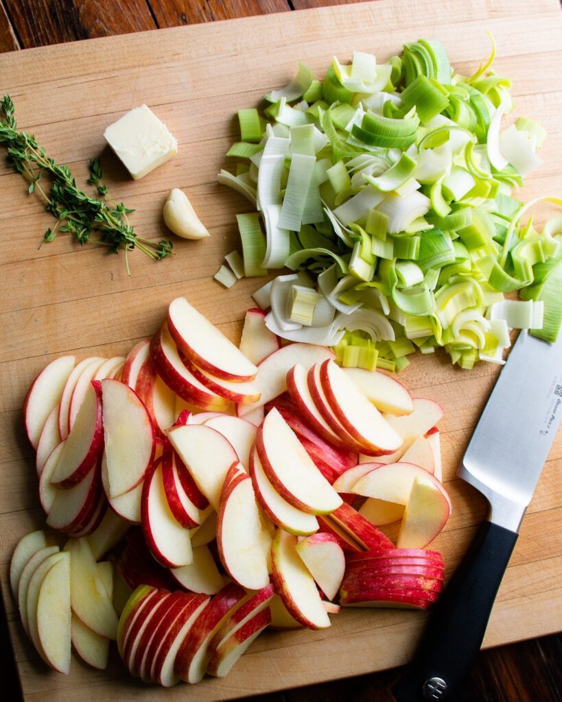 Sliced apples, sliced leeks, butter, garlic and thyme on a wooden cutting board with a chef's knife.