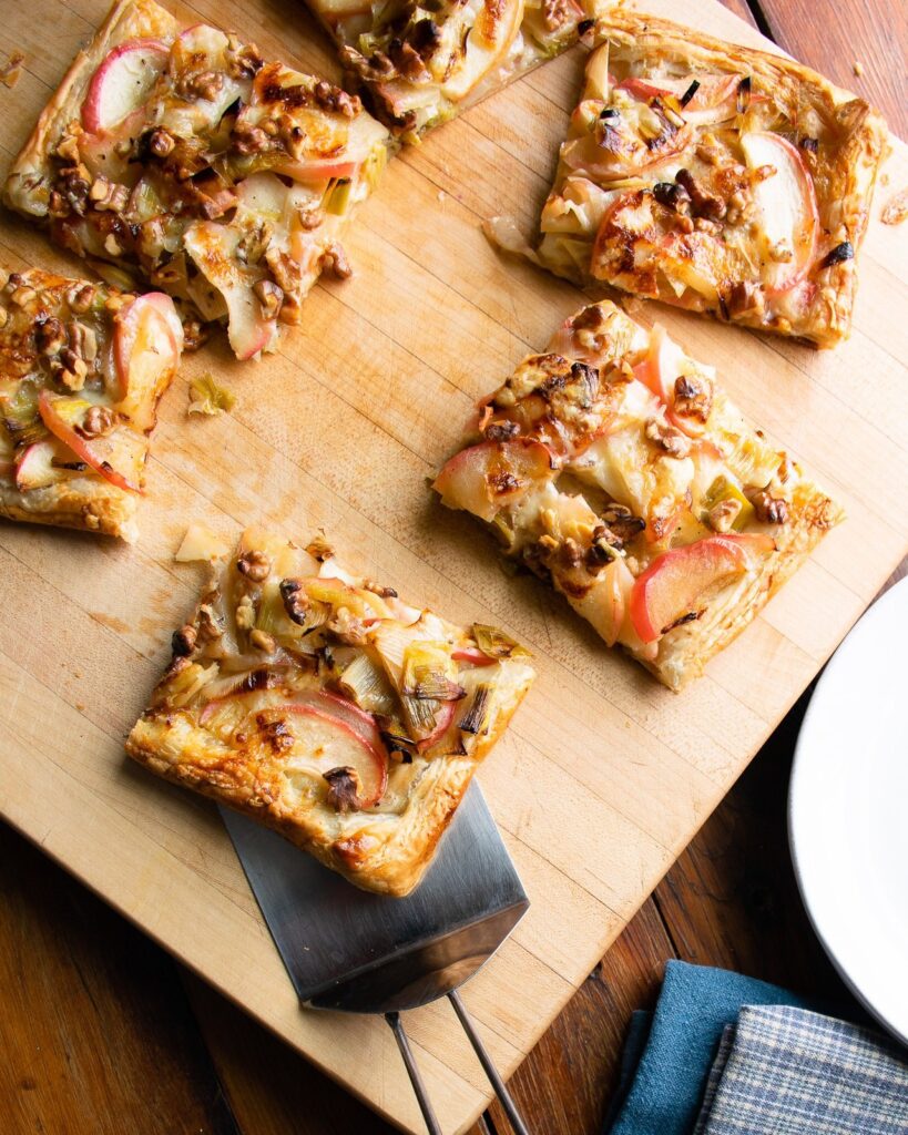 A piece of apple leek cheddar tart on a spatula with the remaining pieces cut up on a wooden cutting board.