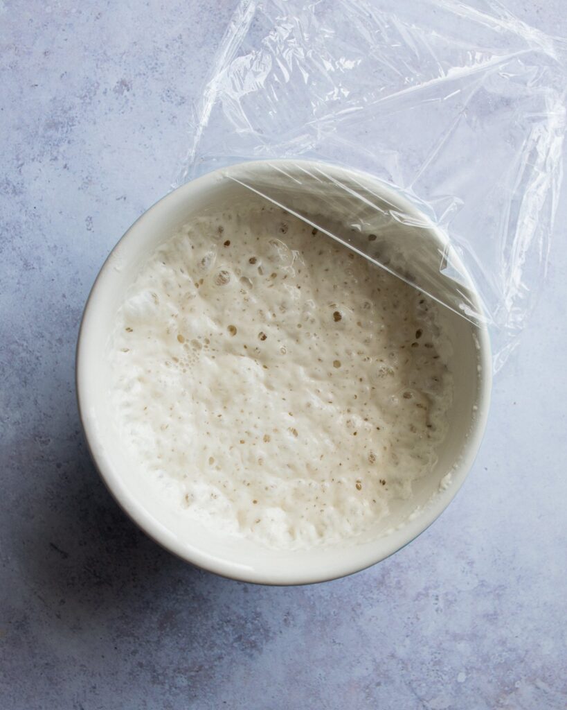 Poolish in a bowl with plastic wrap pulled halfway off the bowl.