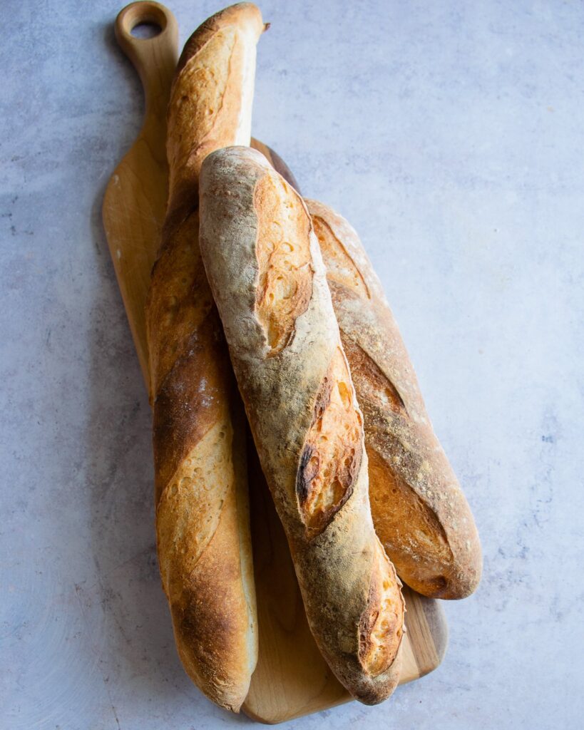 three homemade French baguettes on a wooden board on a counter.