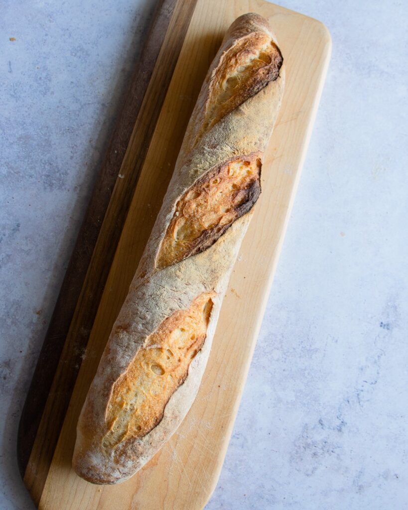 One homemade baguette on a wooden board resting on a marble countertop.