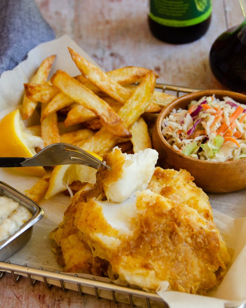 A fork breaking into a beer-battered fish fillet on a parchment lined basket with chips, coleslaw and a beer in the background.