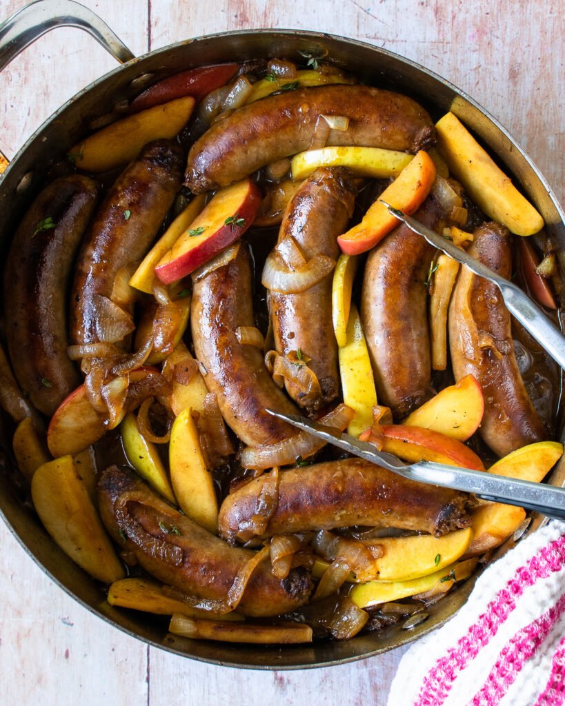 Sausages, onions and apples in a sauté pan with a pair of tongs and a striped towel.