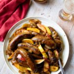Bratwurst with beer, apples and onions on a white plate on a wooden table with a beer and mustard nearby.