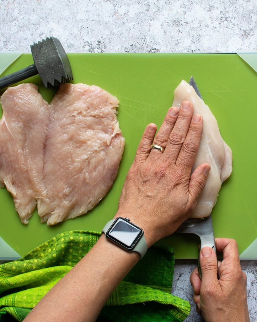 Two hands butterflying a chicken breast on a green cutting board.