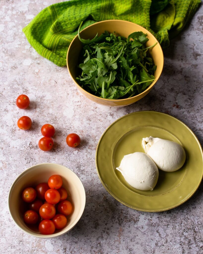 A bowl of arugula, a bowl of cherry tomatoes and a plate with two balls of burrata on a counter.
