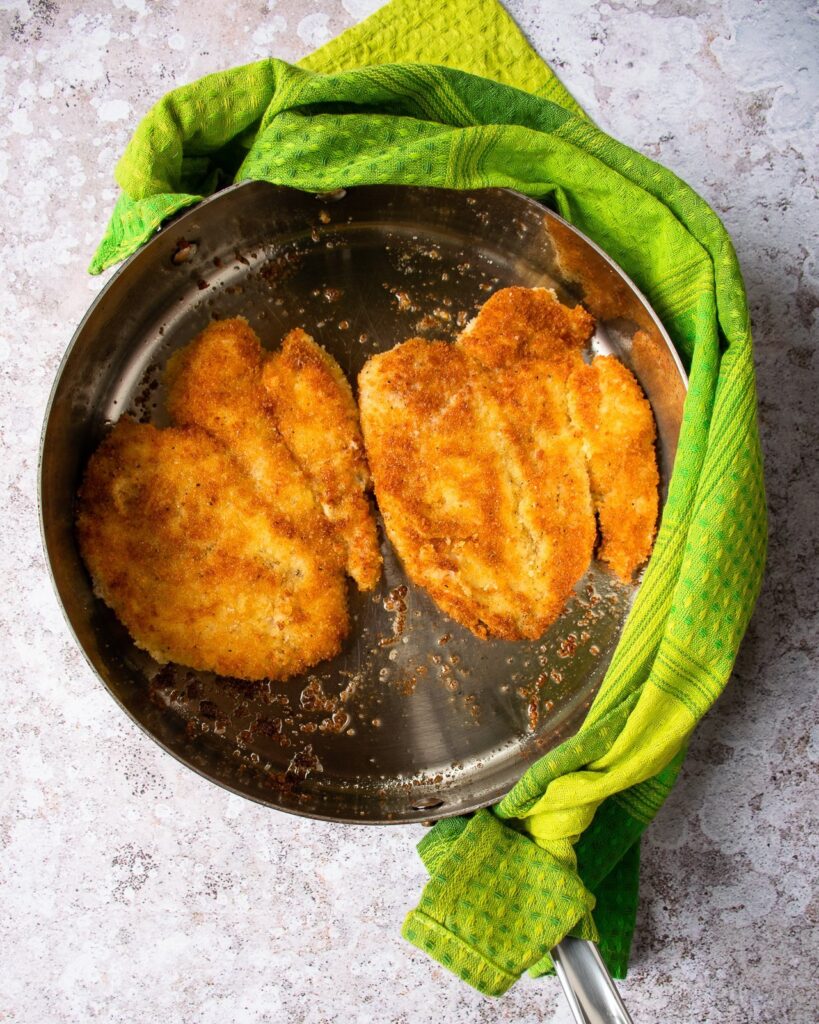 Chicken paillard cooking in a sauce pan with a green towel wrapped around the handle.