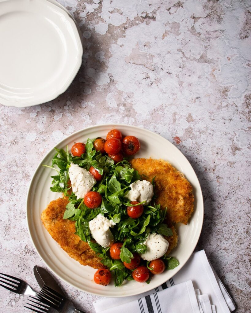 A white plate with chicken paillard, burrata, cherry tomatoes and arugula on a countertop.