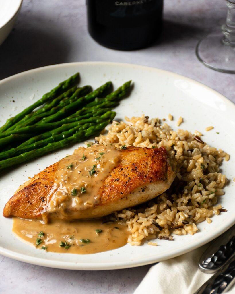 A chicken breast with sherry cream pan sauce, rice and asparagus on a white plate.