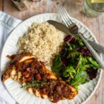 Chicken breasts with tomato balsamic sauce, rice and salad on a white plate with utensils, water, salt and pepper and bread.