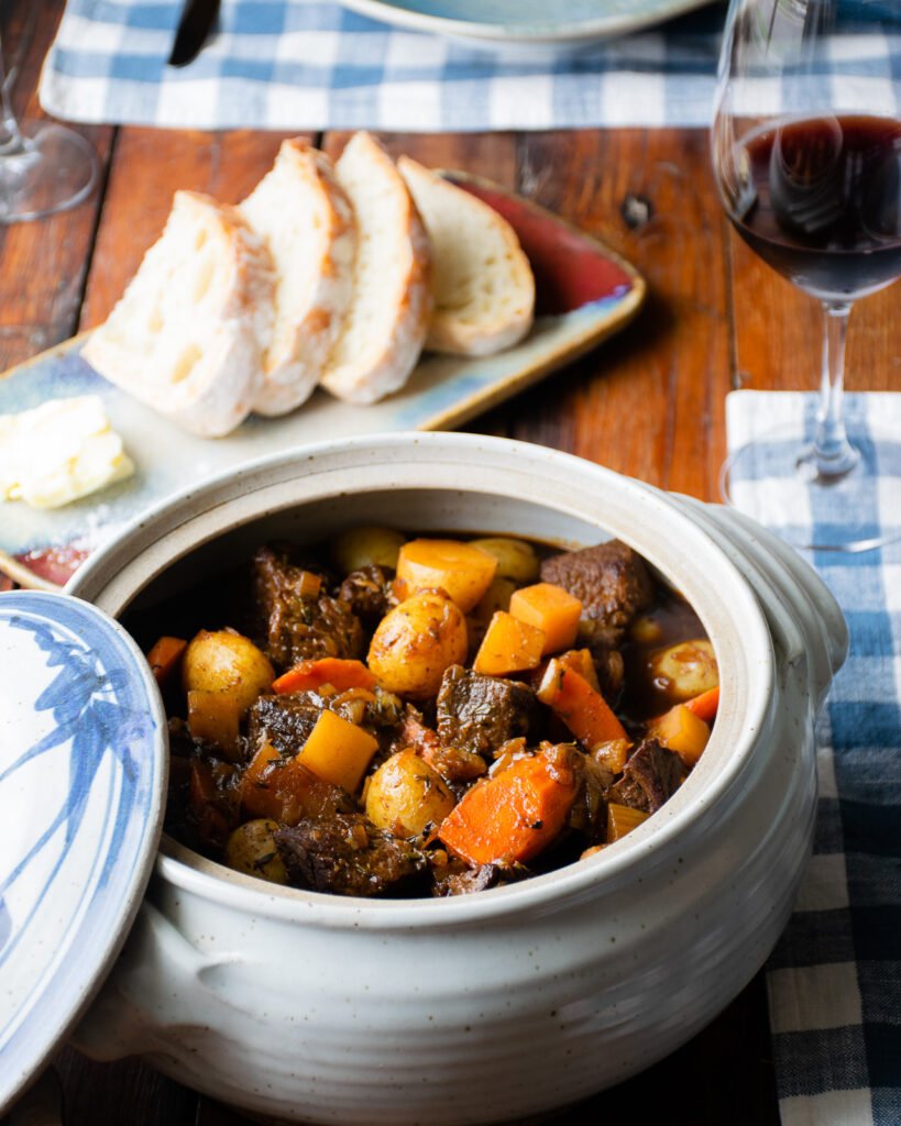 A ceramic casserole filled with Guinness Beef Stew on a table with bread and wine in the background.