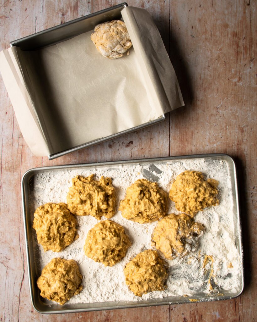 Pumpkin buttermilk biscuit batter in a tray of flour. A square cake pan with one biscuit in it near by.