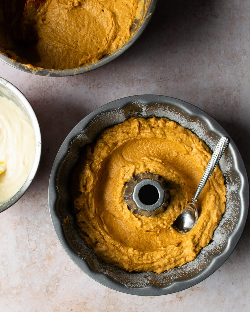 Cake batter in a bundt pan, half full, making a tunnel with the back of a spoon for cream cheese filling.