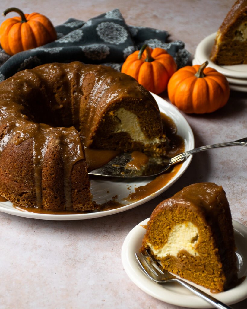 Pumpkin cream cheese bundt cake on a white platter with mini pumpkins in the background and a piece of bundt cake on a small plate in the foreground.