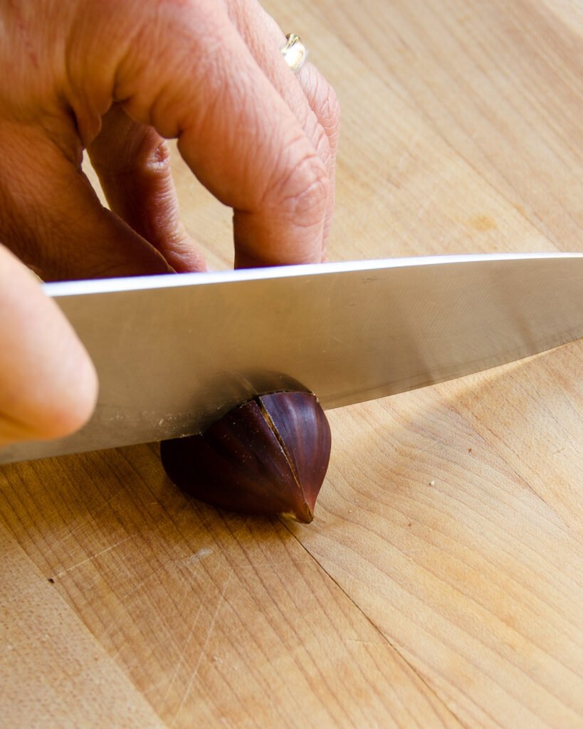 A knife scoring a chestnut on a cutting board.