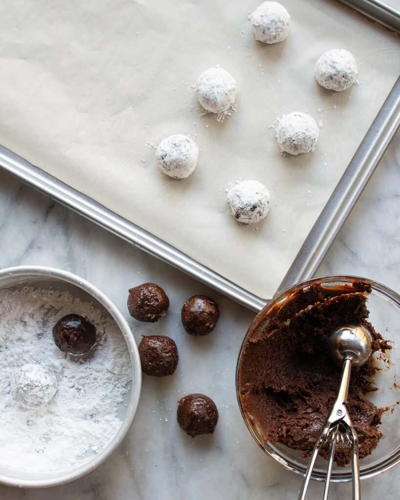 Chocolate Crinkle Cookies in progress - a bowl of dough with an ice cream scoop, a bowl of powdered sugar and some cookies on a baking sheet.