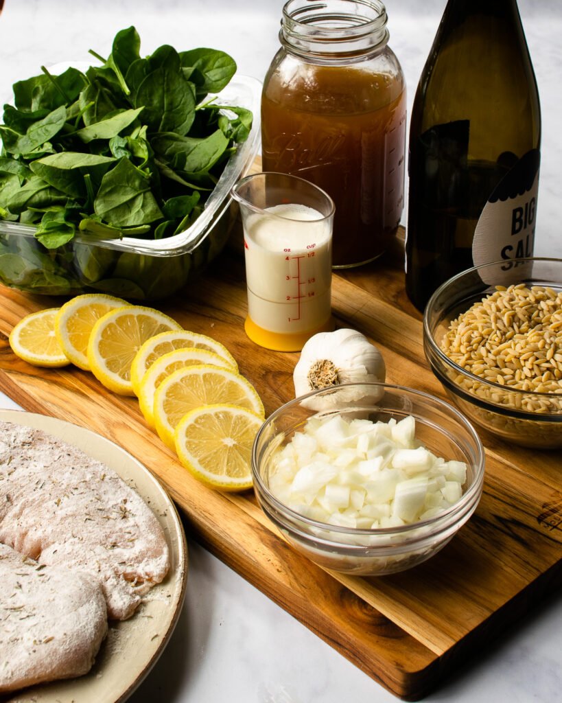 Ingredients on a cutting board - chicken stock, white wine, cream, onion, lemon, chicken, spinach and garlic.