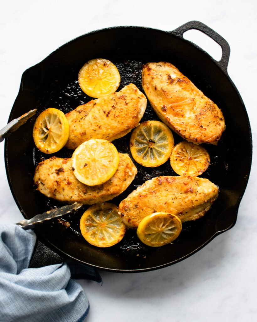 Chicken breasts and lemon slices in a cast iron skillet with tongs and a towel.