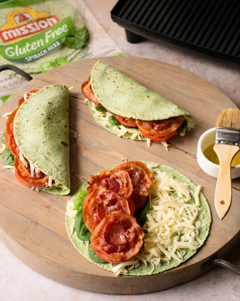 Ingredients for an Italian BLT panini on a cutting board - tortillas, spinach, cheese, tomato.
