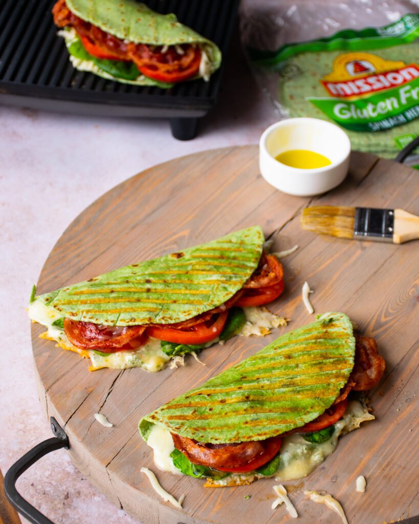 Two grilled paninis on a cutting board with another panini on a panini maker in the background.