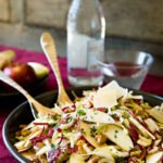 Endive, Radicchio and Apple Salad in a dark bowl with salad servers and a glass bottle of water in the background.