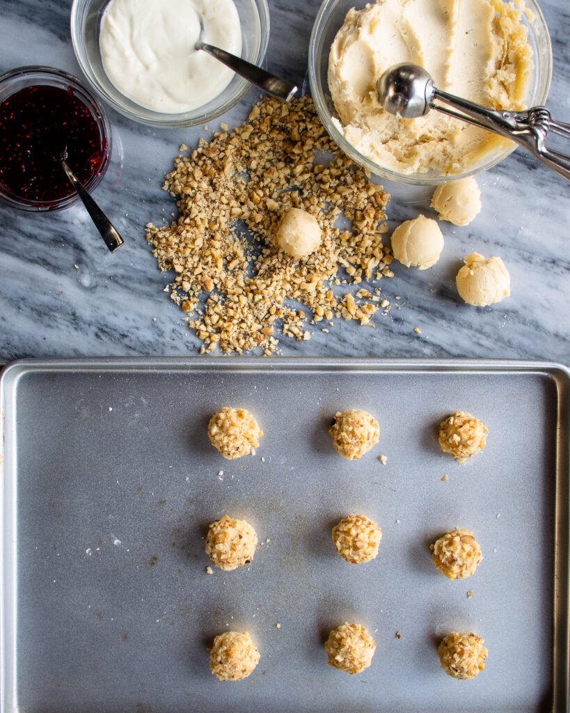 Thumbprint cookies in progress - a bowl of dough, a bowl of cream cheese, a bowl of raspberry jam and some toasted nuts all on a marble countertop with a sheet pan of raw cookie balls.