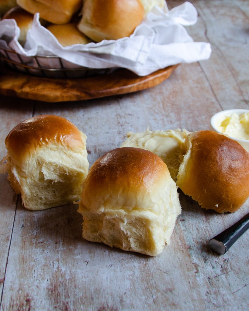 Three bread rolls on a table with knife and butter, with more rolls in aa basket in the background.