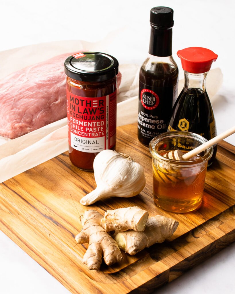 Ingredients on a cutting board - gochujang, soy sauce, sesame oil, honey, garlic and ginger - with a pork loin roast in the background.