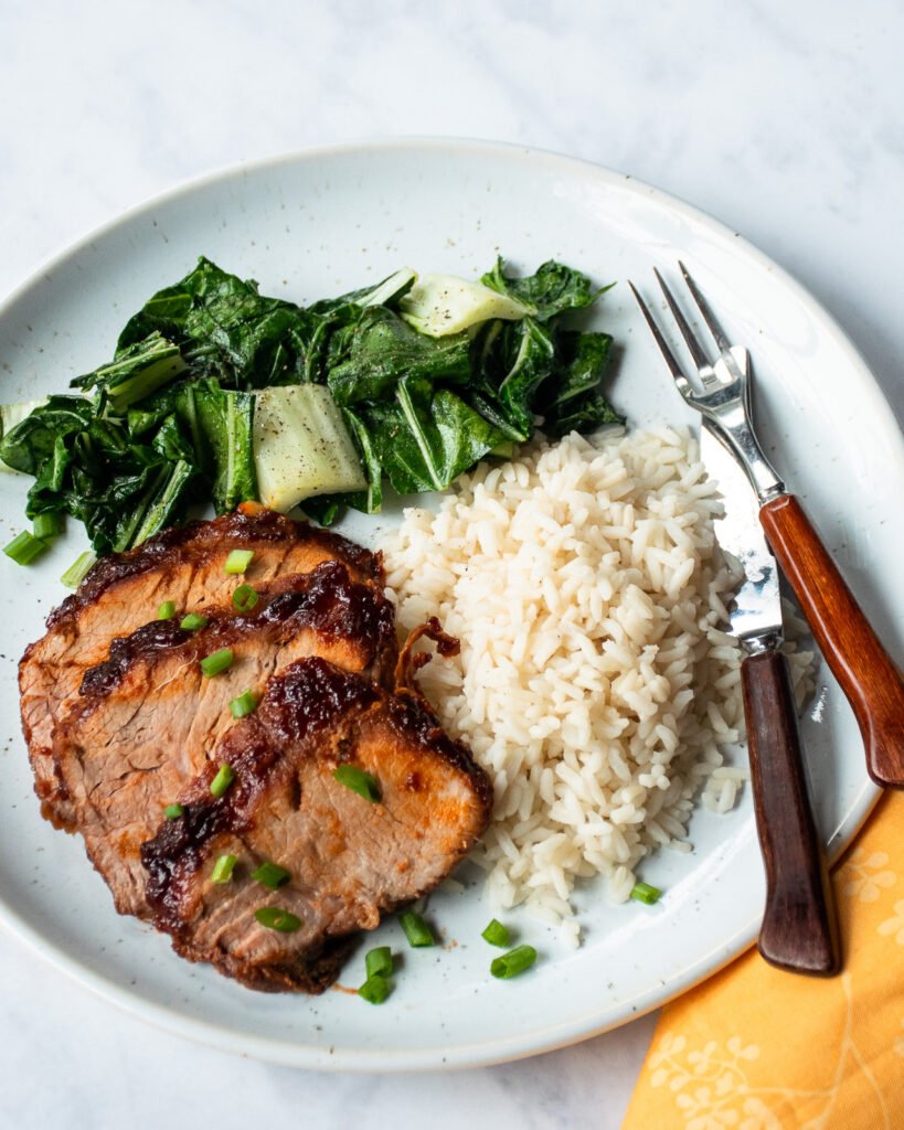A dinner plate with Korean BBQ Pork Loin, white rice, bok choy, knife and fork and a yellow napkin.