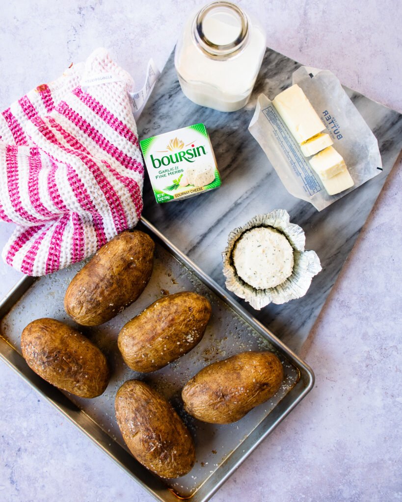 Ingredients on a marble countertop - baked potatoes on a sheet pan, boursin cheese, butter, half and half - and a red and white striped towel.