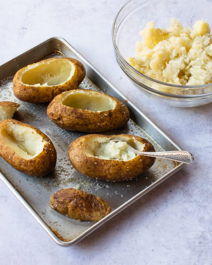 Potatoes on a sheet pan with the tops cut off and the insides scooped out. A bowl of potato in the background.