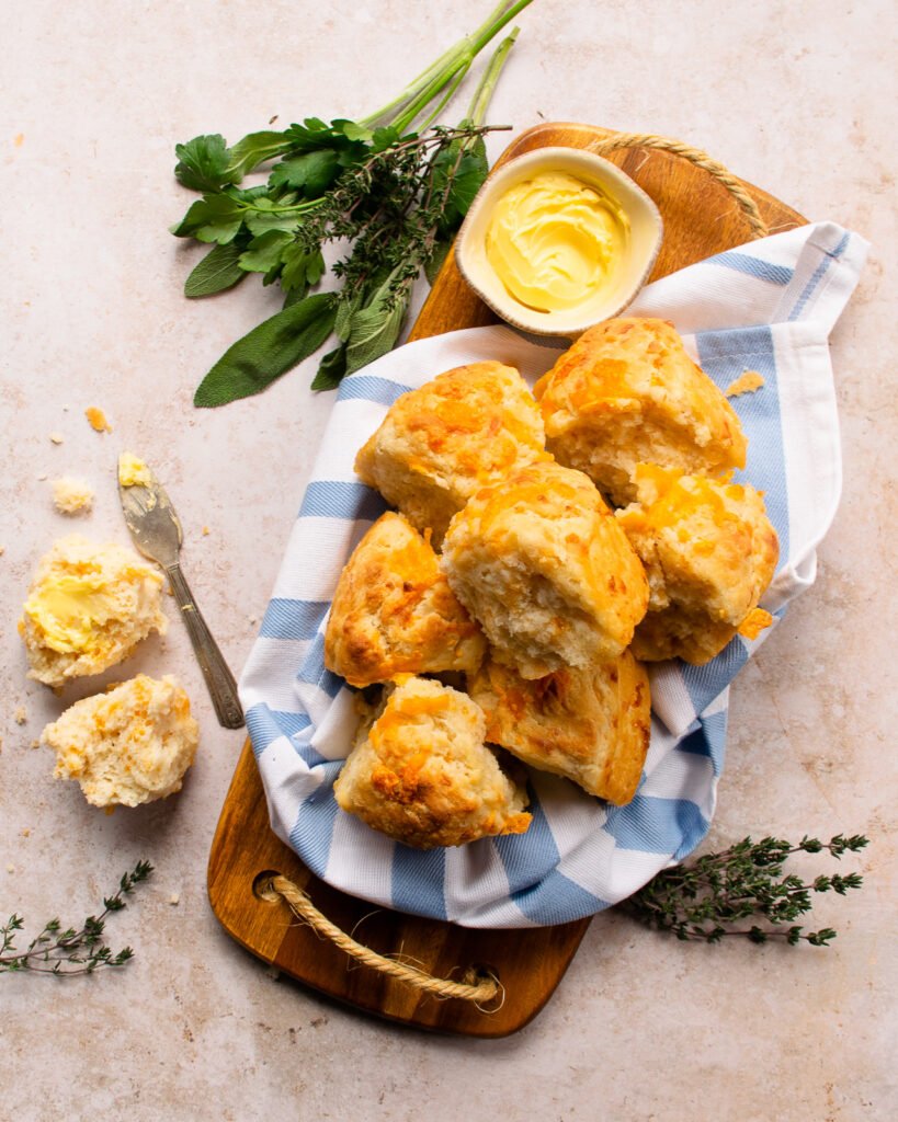Cheddar cheese biscuits on a wooden board with butter and herbs near by. One broken open and buttered.