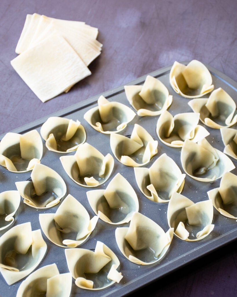 Wonton wrappers, greased and placed into the cups a muffin tin with a stack of wonton wrappers in the background. 