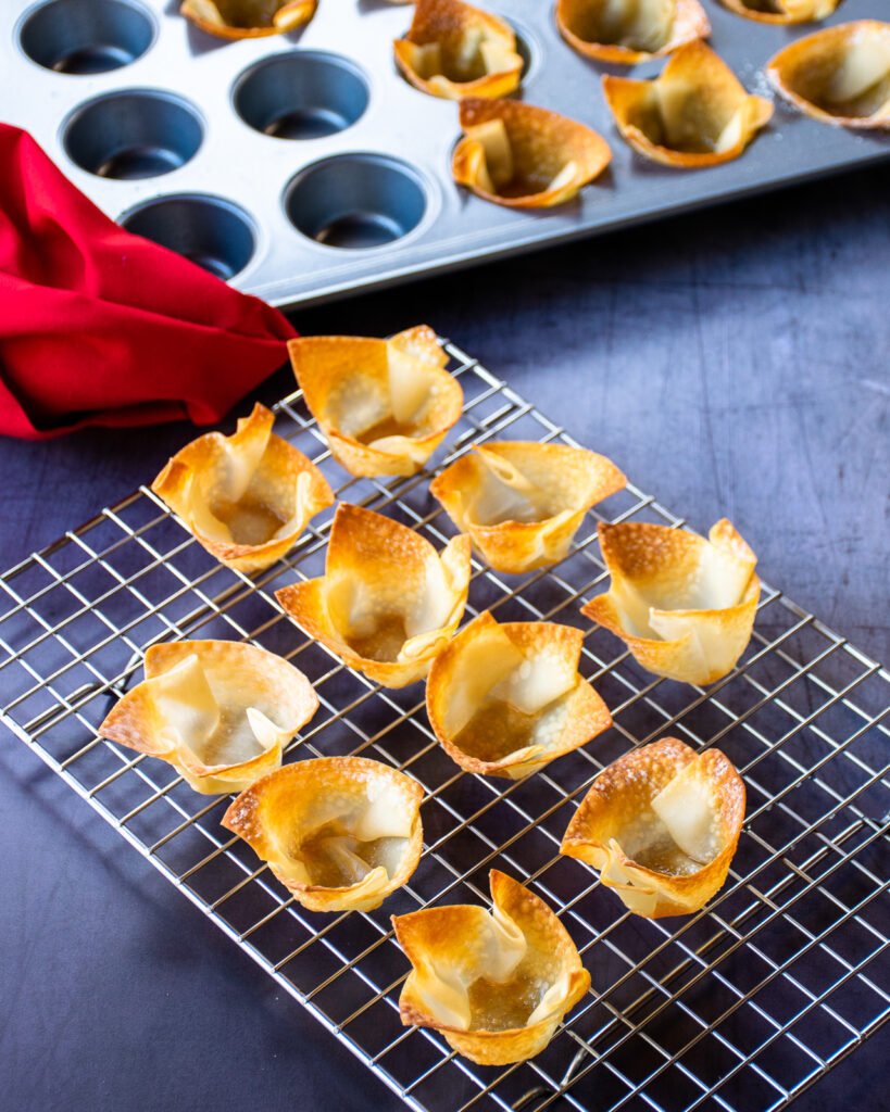 Wonton cups cooling on a cooling rack with a muffin pan in the background and a red napkin.