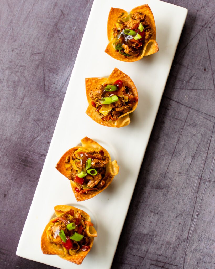 Four chicken egg roll wonton cups on a white serving platter on a concrete counter.