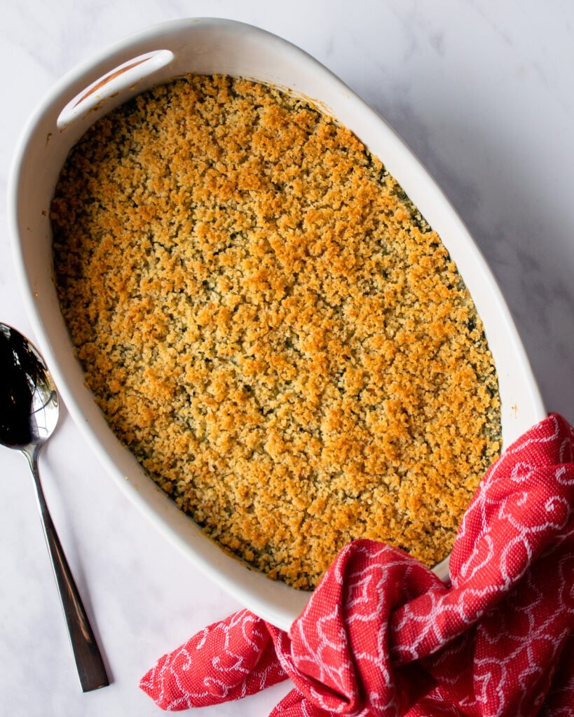 Finished creamed spinach casserole in a white oval ceramic dish with a spoon and red towel