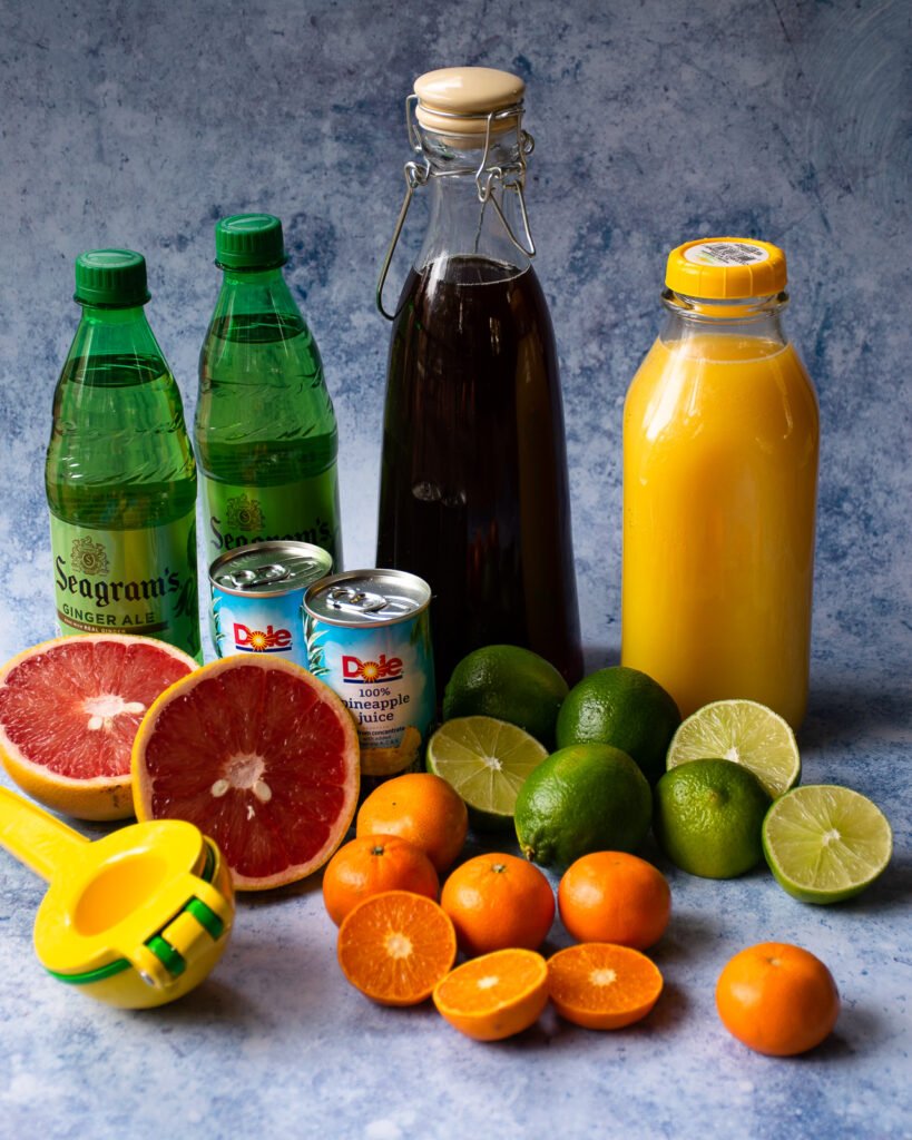 Ingredients for a punch on a blue mottled background - orange juice, tea, ginger ale, grapefruit, mandarins, limes, pineapple juice.