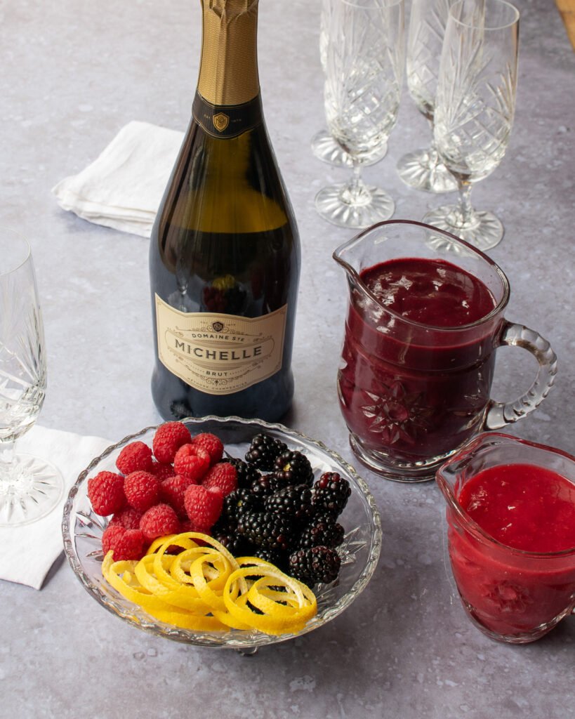 A bottle of prosecco with two cut glass pitchers of fruit puree and a cut glass bowl with raspberries, blackberries and lemon peel garnishes.
