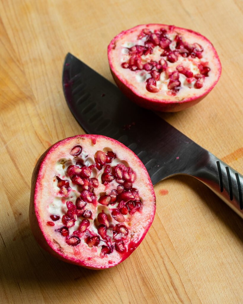 A pomegranate sliced in half on a wooden cutting board with a knife.